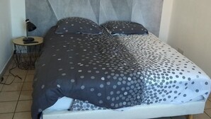 A bed with a grey and white polka dot bedspread, a small bedside table with a lamp, and a plain wall with a geometric pattern. - Studio le Charme d'Ognes (Ognes)