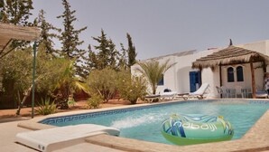 A pool area with lounge chairs, a floating ring, and a building with a thatched roof. - 'DAR Leyla' CHARMING VILLA WITH POOL.... AT THE HEART OF THE DJERBIAN CAMPAIGN (Mezraya)