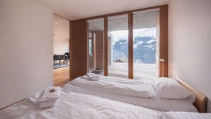A bedroom with two beds, a wooden floor, and a view of mountains through large windows. - Chalet Dillon (Lauterbrunnen)