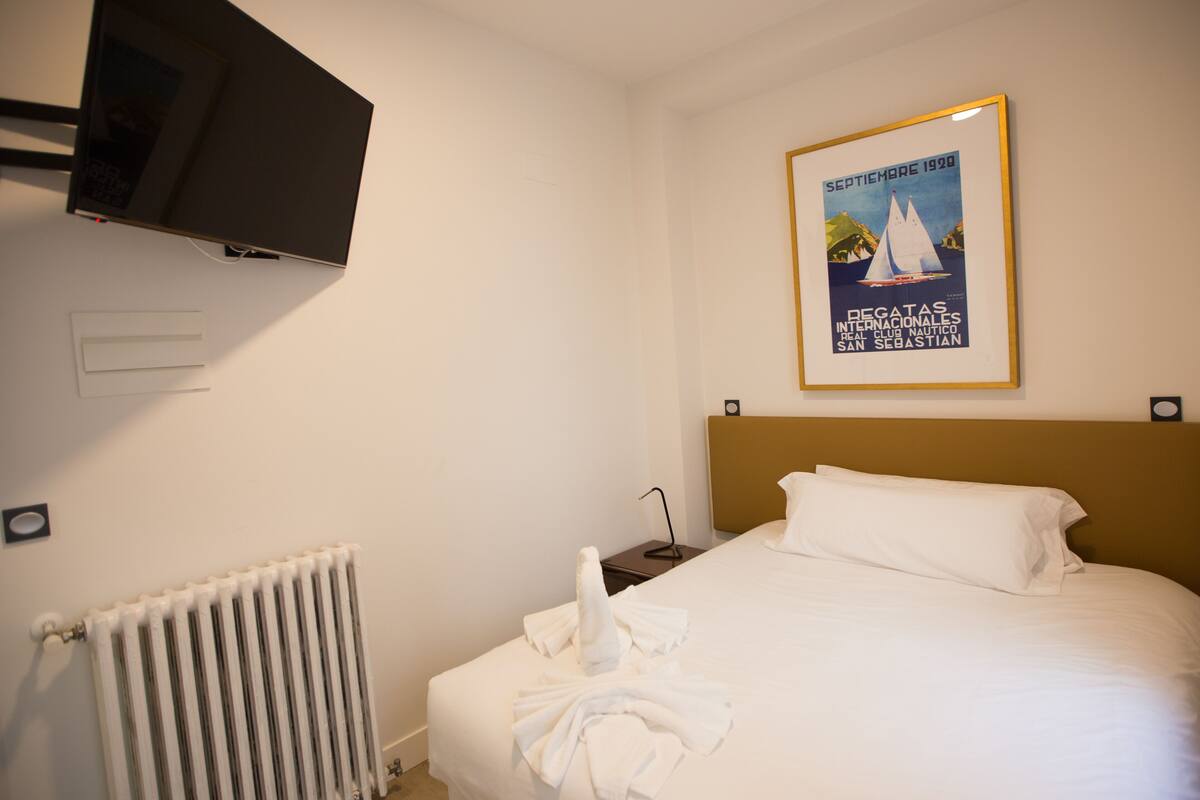 Hotellihuone, jossa on sänky, seinälle asennettu televisio, patteri ja seinällä oleva kehystetty juliste.