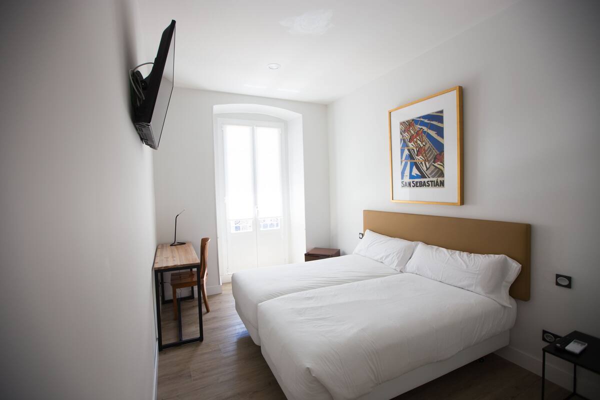 Hotellihuoneessa on suuri sänky, työpöytä ja tuoli. Huoneessa on ikkuna, josta avautuu näkymä ulkona olevaan rakennukseen.