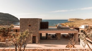 A modern villa with a stone facade, a covered outdoor seating area, and a view of the sea.