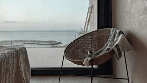 Een rieten stoel bij een groot raam met uitzicht op een rustige zee en rotsformaties.