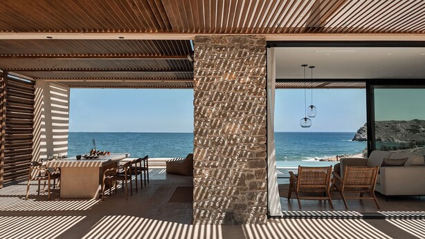 Een modern terras met uitzicht op de zee, met een stenen pilaar, rieten meubels en een heldere lucht.