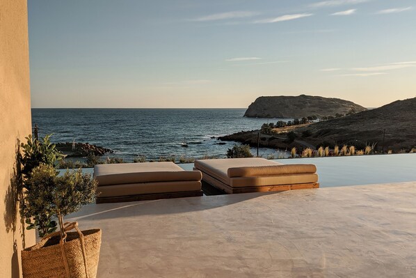 An infinity pool with beige loungers, a small potted plant, and a view of the sea and a rocky hill.