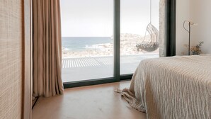 Een slaapkamer met een groot raam met uitzicht op een strand, een hangstoel en een bed met een geribbelde deken.
