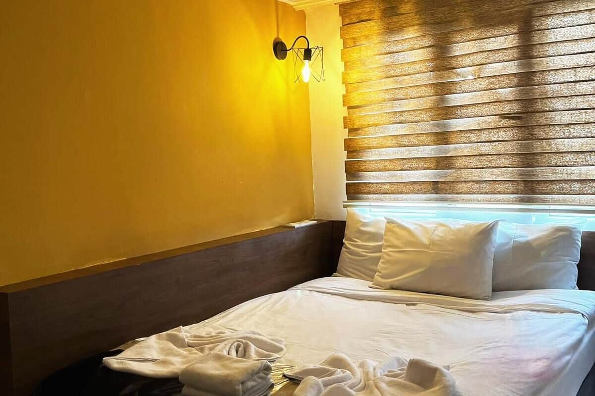 Ein Hotelzimmer mit Bett, Nachttisch, Wandlampe und Fenster mit Jalousien.
