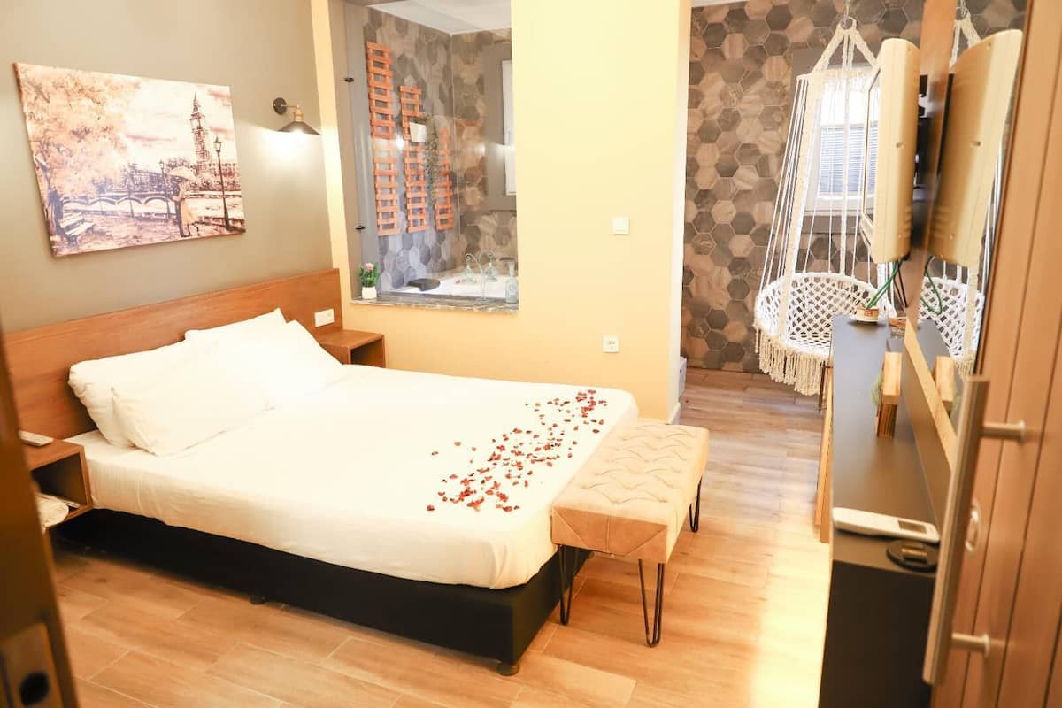Ein Hotelzimmer mit einem großen Bett, einer Holzbank, einem kleinen Tisch, einem Hängekorbstuhl und Blick auf eine Brücke und Gebäude durch ein Fenster.