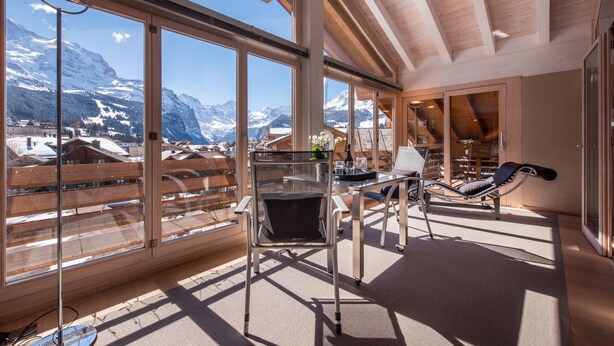 A spacious balcony with large windows offering a mountain view, a comfortable lounge chair, and a table with a vase of flowers.