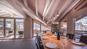 A dining area with a long wooden table set for a meal, large windows offering a view of the mountains, and a high ceiling with exposed beams.