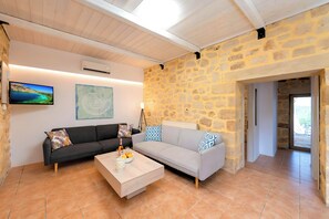 A living room with a stone wall, a grey sofa, a black sofa, a wooden coffee table, and a television. - Cretan Breeze Villa | Heated Pool (Georgioupoli)