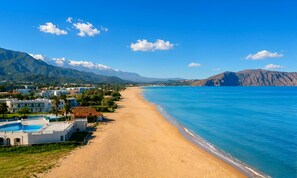 A beach with a pool, buildings, and mountains in the background.