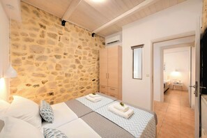 A bedroom with a stone wall, wooden ceiling, a bed with a patterned bedspread, a bedside table with a lamp, and a wardrobe. - Cretan Breeze Villa | Heated Pool (Georgioupoli)