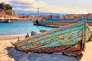 A weathered boat with the inscription "NOYNOY" on a sandy beach, with a coastal town and mountains in the background. - Cretan Breeze Villa | Heated Pool (Georgioupoli)