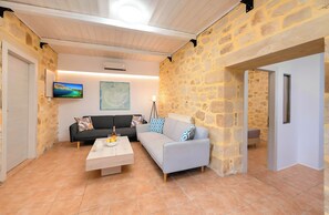 A cozy living room with stone walls, a grey sofa, a white coffee table, and a TV mounted on the wall. - Cretan Breeze Villa | Heated Pool (Georgioupoli)