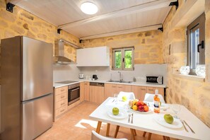 A modern kitchen with a stone wall, stainless steel appliances, and a dining area set for two with fruit and wine. - Cretan Breeze Villa | Heated Pool (Georgioupoli)