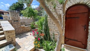 A stone-walled courtyard with a wooden door, steps leading to a covered area, and various plants including flowers and a cactus.