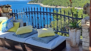 A balcony with wicker furniture, a stone wall, and a view of the sea and green hills.