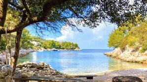 A bench by a clear, calm body of water with rocky shores and trees in the background.