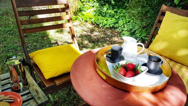 A wooden chair with a yellow cushion, a round table with a tray containing a pitcher, cups, and strawberries, and a metal teapot on a wooden bench. - At home in the love nest (Innsbruck)