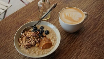 Un tazón de avena con arándanos y granola, una taza de café con arte de latte, y una botella pequeña de aceite sobre una mesa de madera.