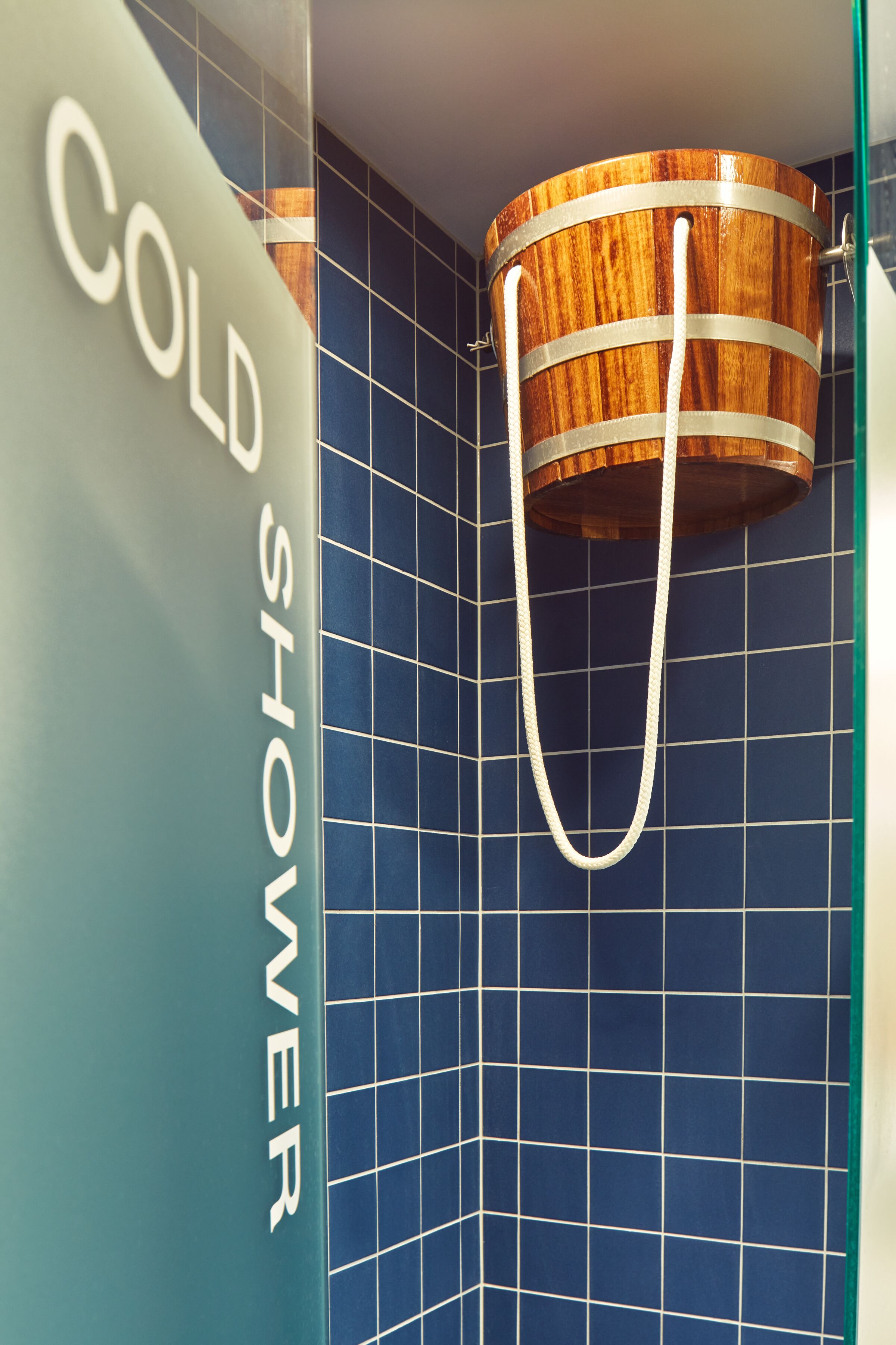 Un baño con pared de azulejos azules, un cubo de madera colgado de un gancho metálico y un letrero que dice 'COLD SHOWER'.