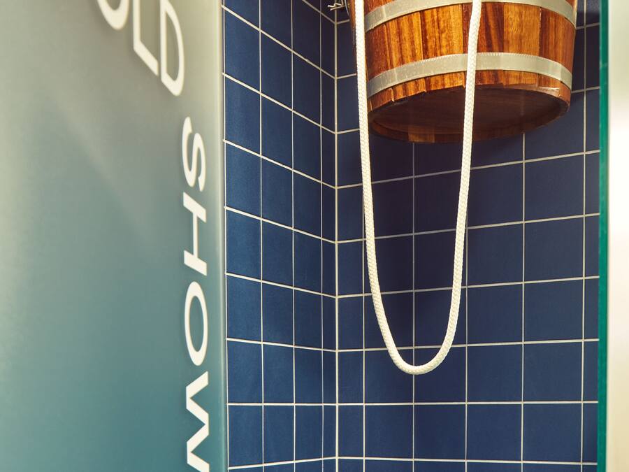 Un baño con pared de azulejos azules, un cubo de madera colgado de un gancho metálico y un letrero que dice 'COLD SHOWER'.