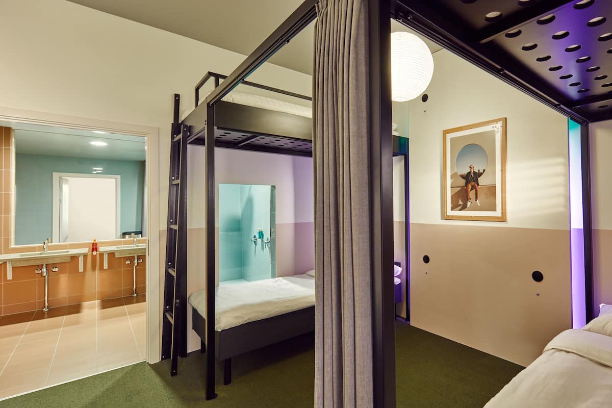 Una habitación de hotel moderna con una cama litera, un baño con mampara de vidrio y un espejo grande.