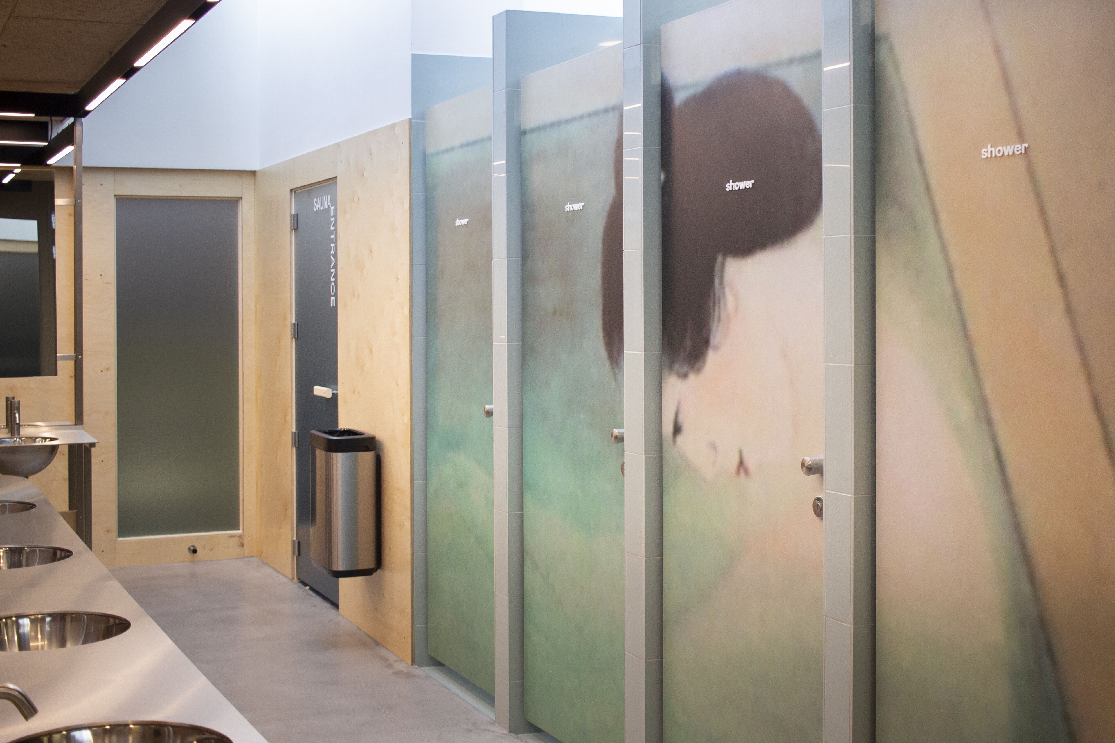 Baño público con varios lavamanos, un cubo de basura y mamparas de vidrio esmerilado.