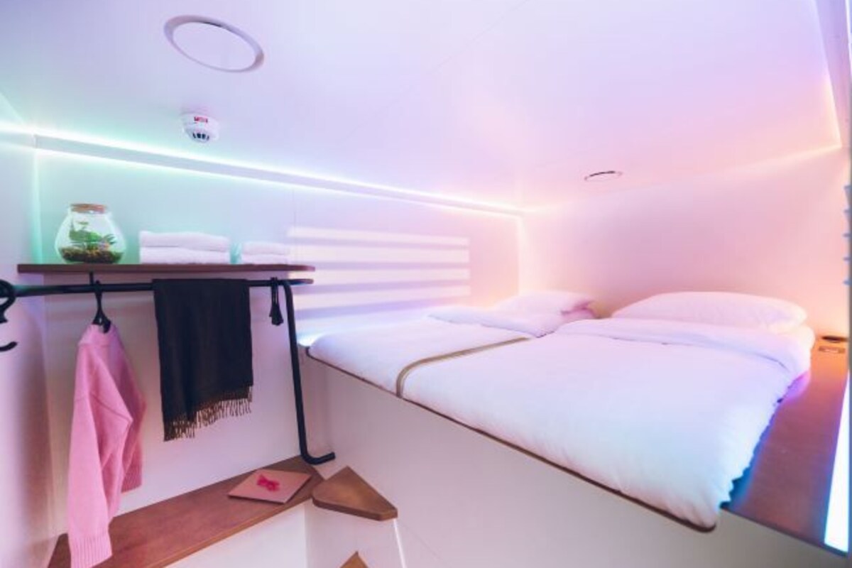 Una habitación de hotel moderna con una cama, una mesita con toallas y una estantería con un objeto decorativo.