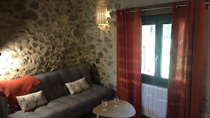 A cozy room with a stone wall, a grey sofa with patterned cushions, a small round table with wine glasses, and red curtains. - Charming stone house in the countryside (Céret)