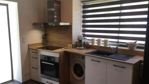 A compact kitchen with a built-in oven, microwave, and washing machine. It features a window with blinds and a stainless steel range hood. - Charming stone house in the countryside (Céret)