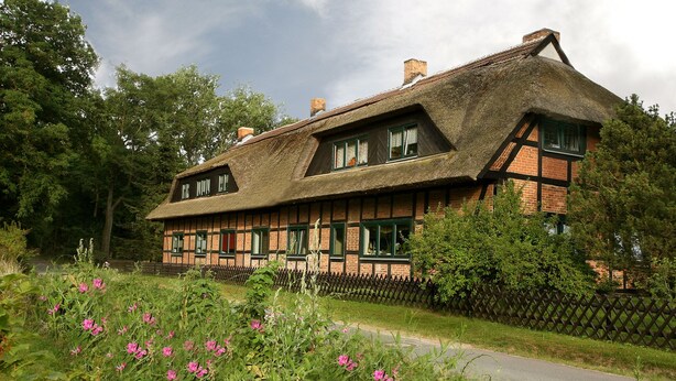 Ein traditionelles Fachwerkhäuschen mit Strohdach, umgeben von einem Holzzaun und blühenden Pflanzen.