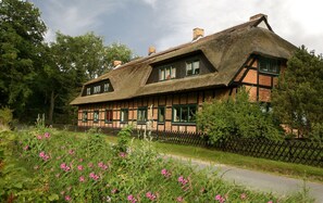 Ett traditionellt hus med korsvirkesfasad och halmtak, omgivet av ett trästaket och blommande växter.