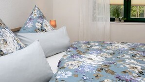Ein Schlafzimmer mit blumigem Bettbezug, weißen Kissen, einer Nachttischlampe und einem Fenster mit Blick ins Grüne.