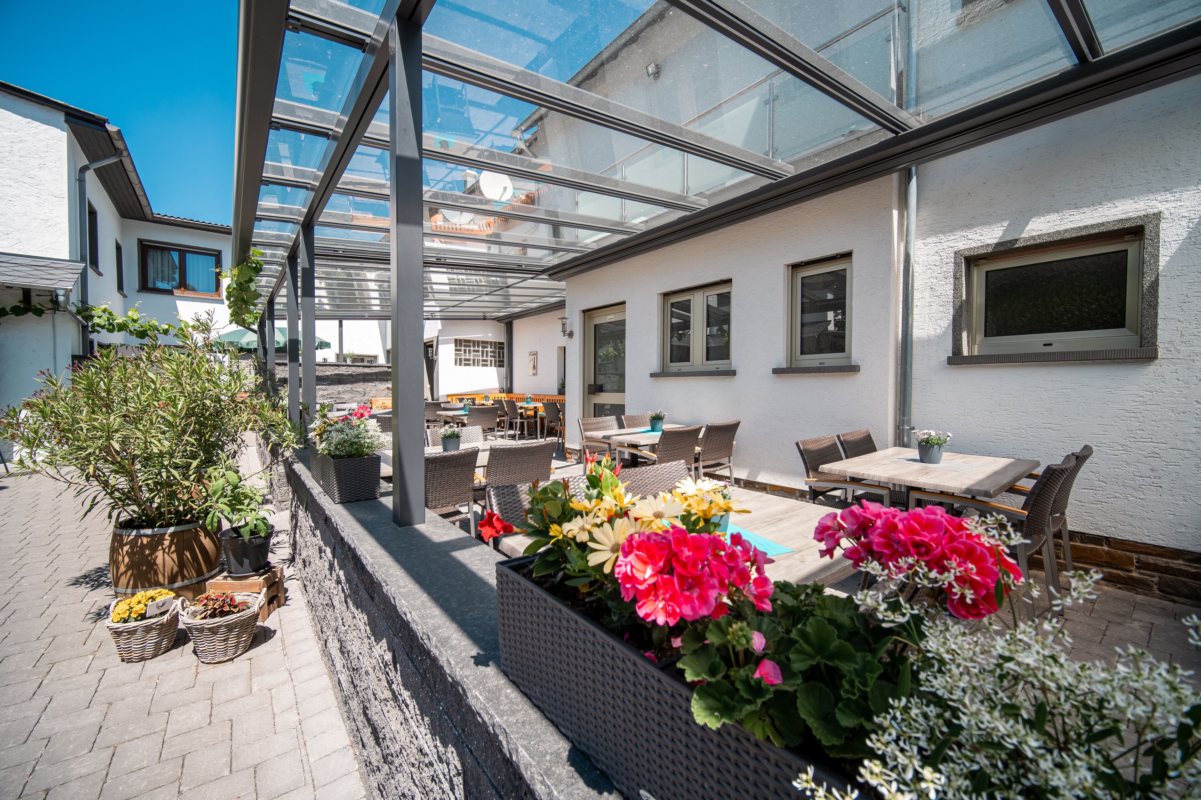 Eine überdachte Terrasse im Freien mit Tischen, Stühlen und Topfpflanzen, sowie einer Glasüberdachung.