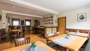 A spacious dining area with wooden tables and chairs, a kitchen in the background, and framed pictures on the walls. - Hotel Langen (Loef)