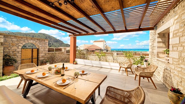 Zona d'exterior per a dinar amb taula de fusta preparada per a quatre persones, rodejada de cadires de vimet i vistes panoràmiques al mar i a les muntanyes.