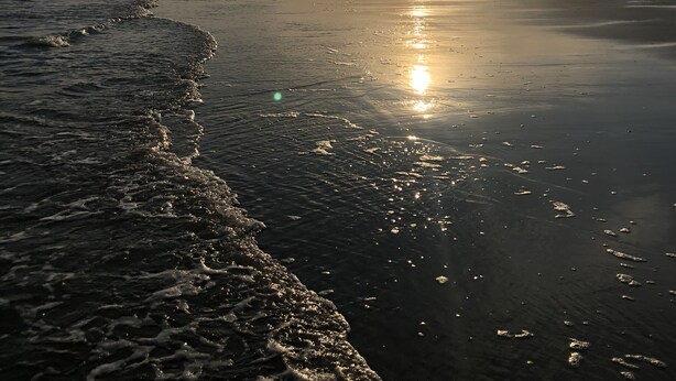 Een strand met de zon die ondergaat aan de horizon, golven spoelen zachtjes op het strand.