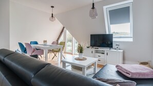 Een moderne woonkamer met een zwarte leren bank, een witte salontafel met een plant, een televisie op een witte kast, en een eethoek met kleurrijke stoelen.