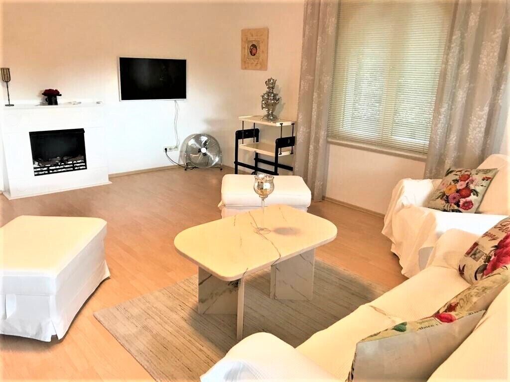 Ein Wohnzimmer mit einem weißen Sofa, einem kleinen weißen Tisch, einem Kamin, einem an der Wand montierten Fernseher und einem fenster mit Vorhängen.