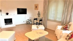 En stue med en hvid sofa, et lille hvidt bord, en pejs, et tv ophængt på væggen og et vindue med gardiner.