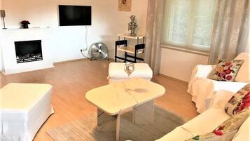 Ein Wohnzimmer mit einem weißen Sofa, einem kleinen weißen Tisch, einem Kamin, einem an der Wand montierten Fernseher und einem fenster mit Vorhängen.