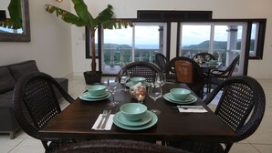A dining area with a dark wooden table set for four, surrounded by wicker chairs, with a large window offering a view of greenery and mountains. - Beautiful supersize villa perfect for larger families and groups. Sleeps 22 (Cap Estate)