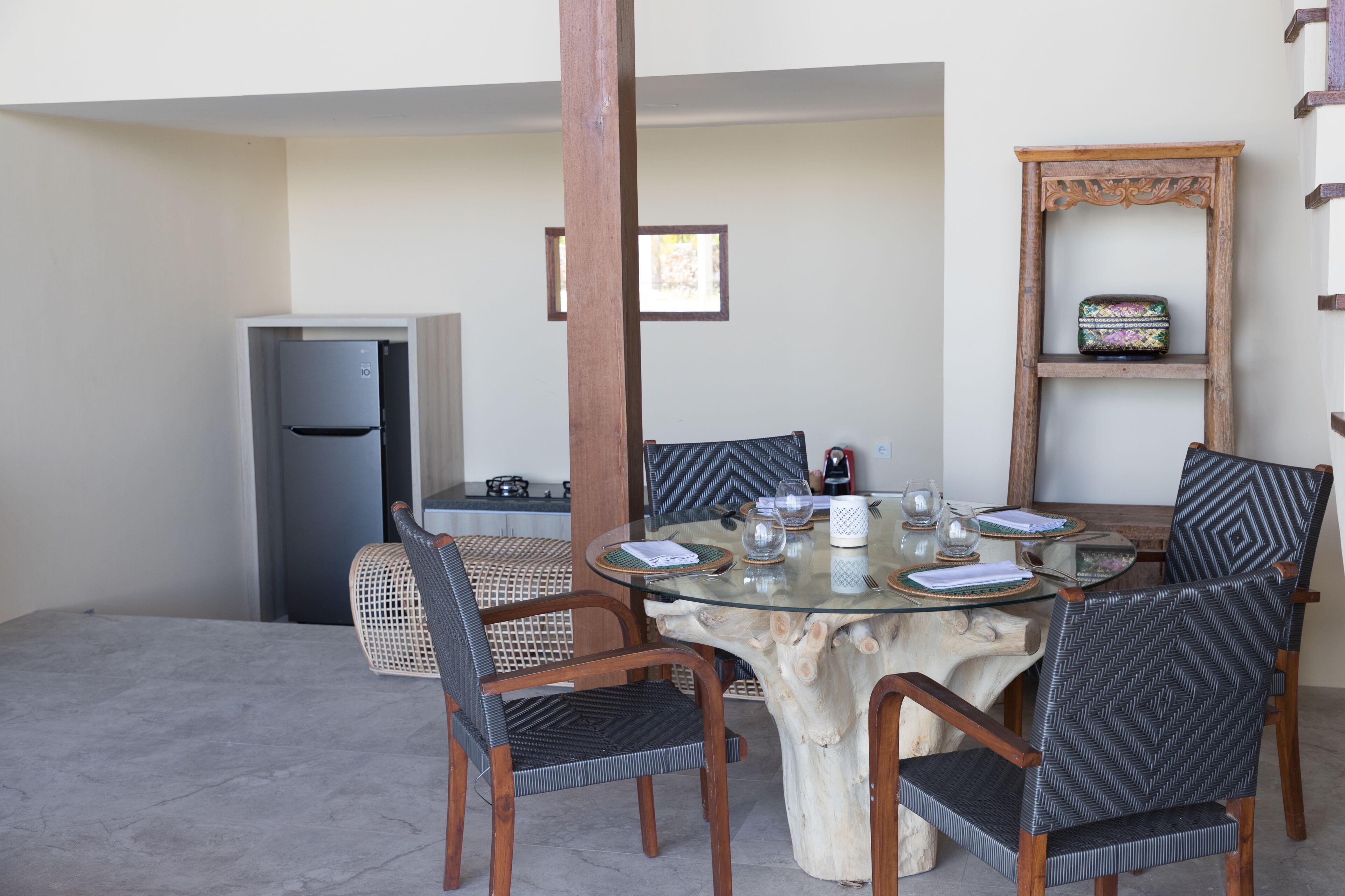 A dining area with a round glass table surrounded by chairs, a wooden chair, a small refrigerator, and a decorative shelf.