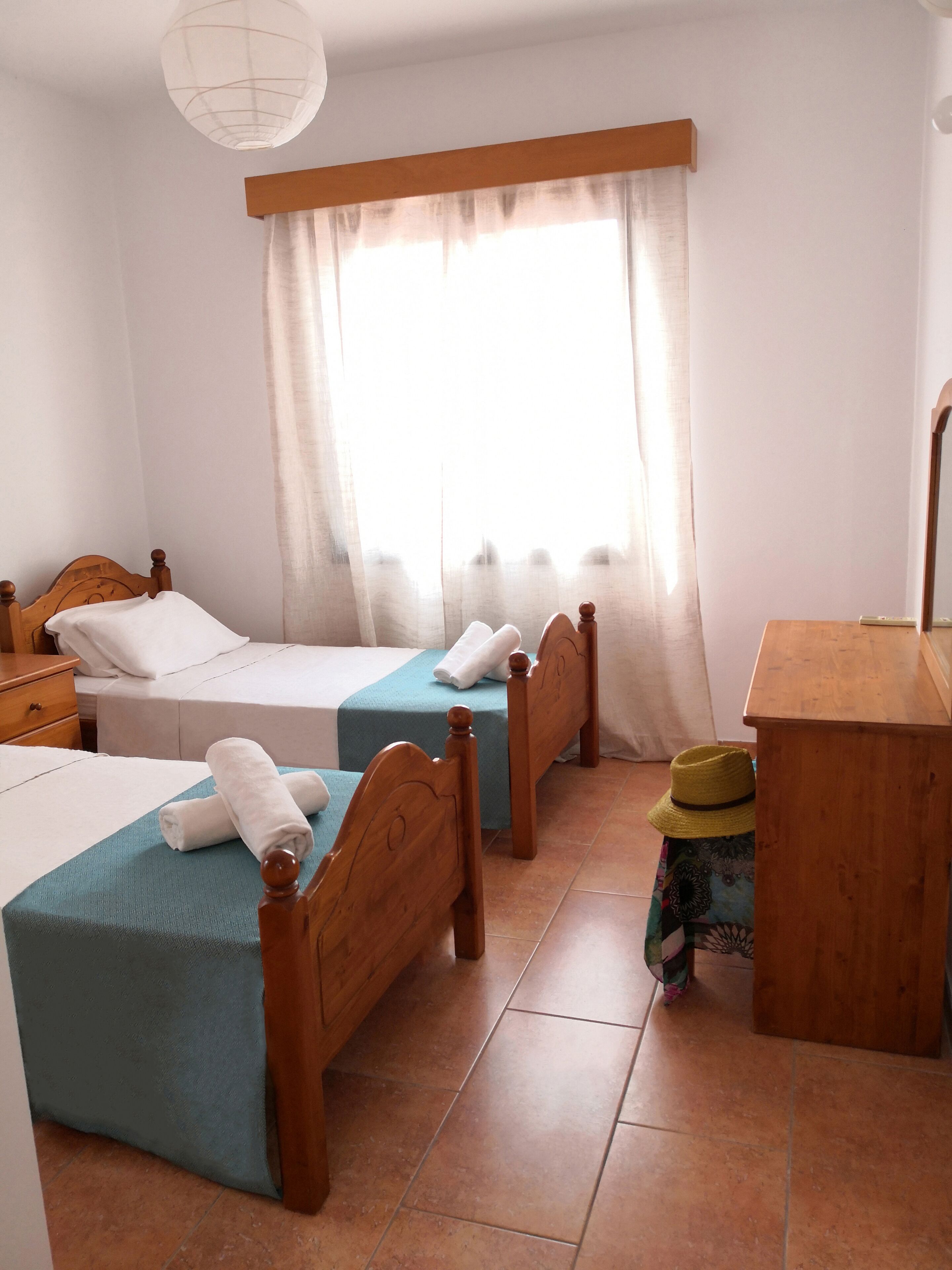Camera d'albergo con due letti singoli, una scrivania in legno, uno sgabello e un'ampia finestra con tende.