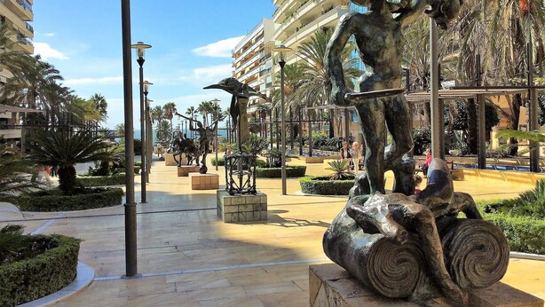 A plaza with sculptures, palm trees, and modern buildings.