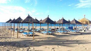 A beach with rows of sun loungers and thatched umbrellas, a clear sky, and the ocean in the background.