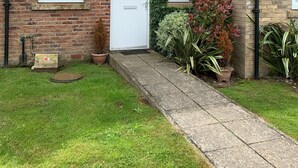 Une maison à deux étages, construite en briques, avec une porte d’entrée blanche, un petit porche et un jardin agrémenté de plantes en pot.