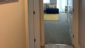 Un couloir mène à une chambre qui contient un lit, une table de chevet et une fenêtre avec des rideaux. Une pancarte indiquant « Bienvenue chez nous » est accrochée au-dessus de la porte.